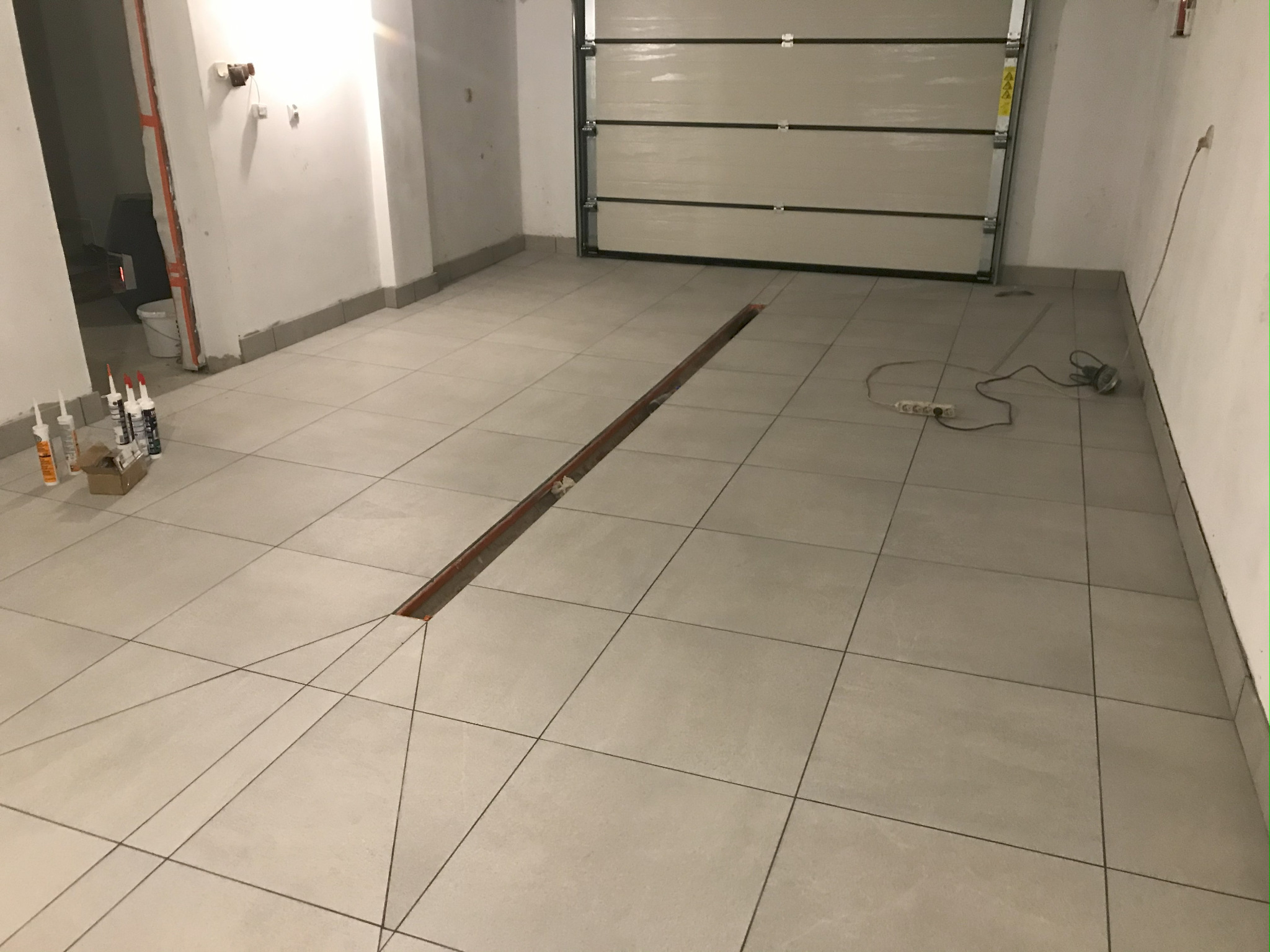 Jasne płytki podłogowe w garażu z odpływem liniowym. Tuby z klejem i przedłużacz elektryczny w tle. Widok na bramę garażową i fragment ściany.