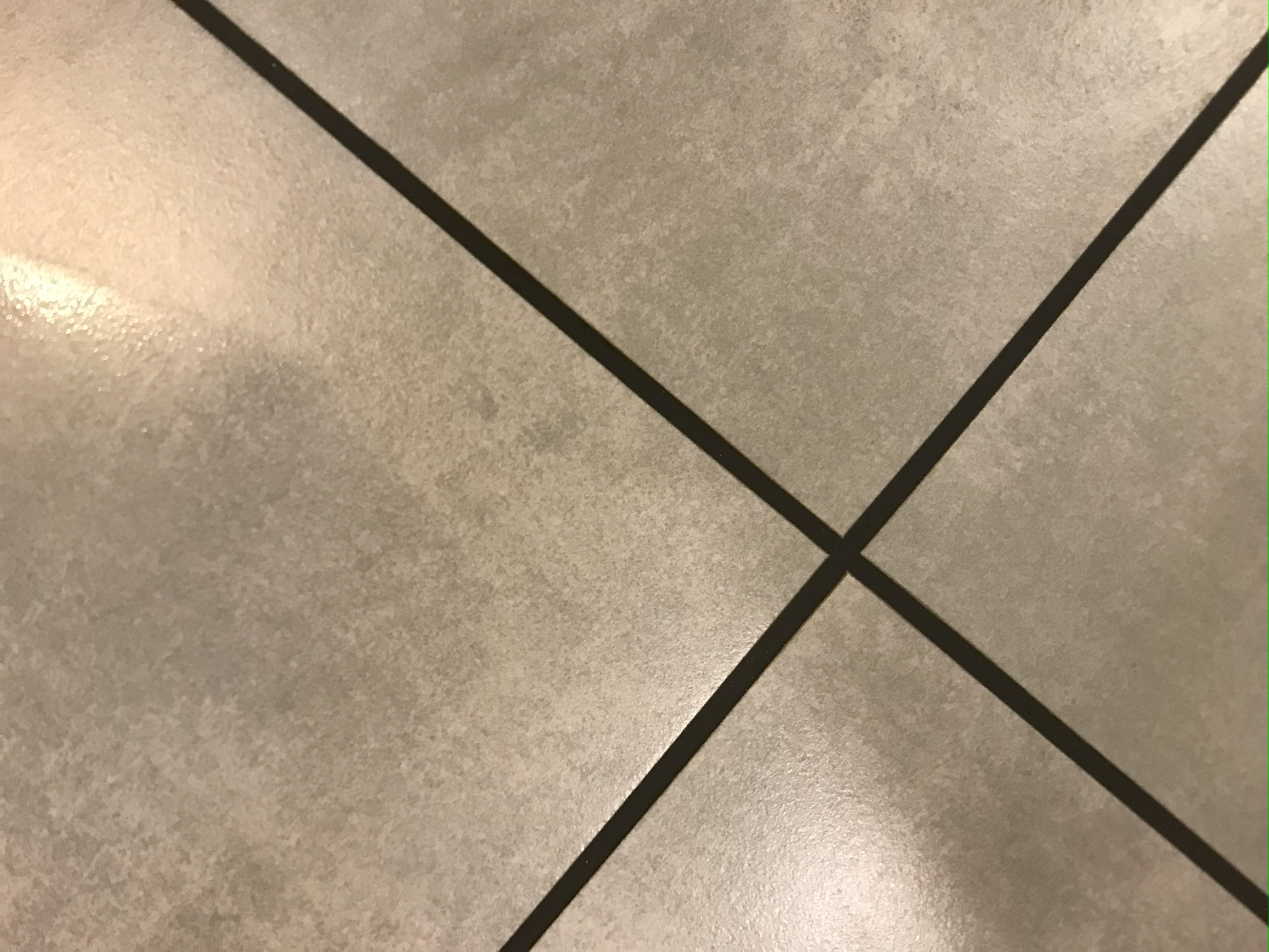 Szare, kwadratowe płytki podłogowe z ciemną fugą, ułożone pod kątem, tworzące wzór krzyża w miejscu przecięcia. Widoczne delikatne refleksy światła na powierzchni.