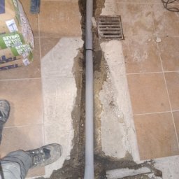 Sebastian Gajewski - Szara rura kanalizacyjna w wykopanym bruzdzie w podłodze z płytek, widoczna krata ściekowa, worek z betonem i but. Etap prac podczas instalacji.