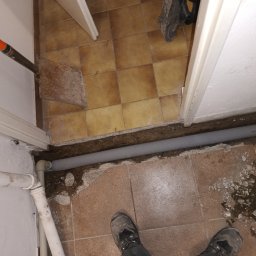 Sebastian Gajewski - Szara rura kanalizacyjna w wykopanym rowku w podłodze z płytek, widoczne narzędzie i fragment buta. Prace hydrauliczne w trakcie remontu łazienki.