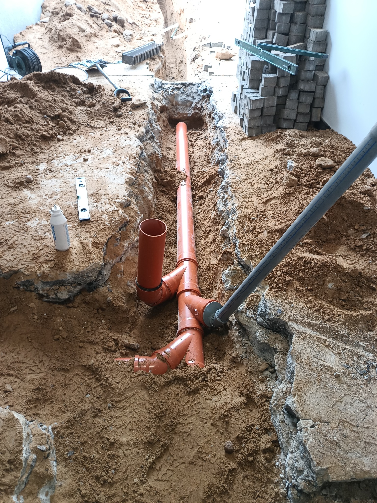 Wykonana instalacja kanalizacyjna z pomarańczowych rur w wykopie ziemnym, obok ułożona kostka brukowa. Widoczny poziomica i narzędzia.