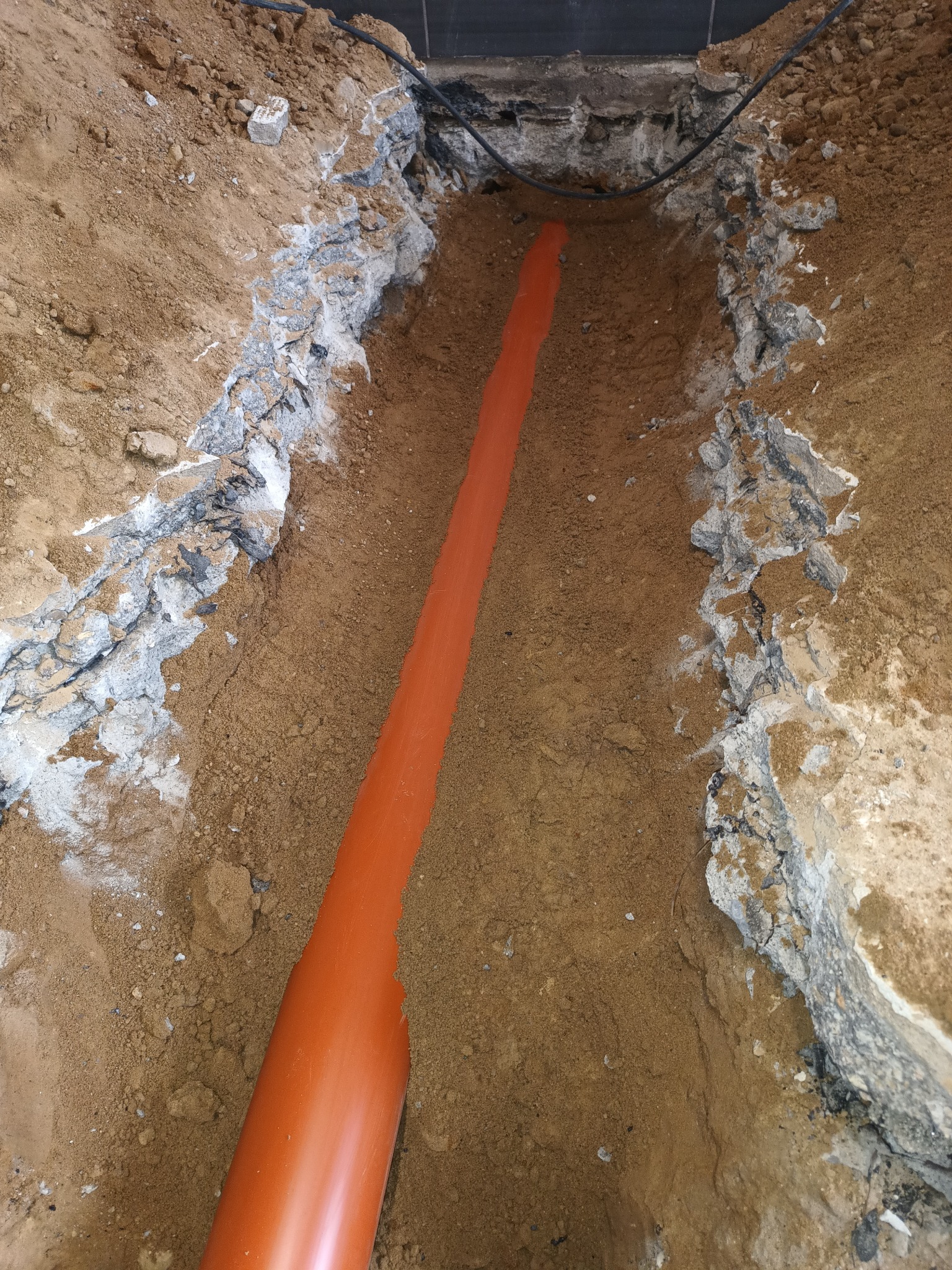 Pomarańczowa rura kanalizacyjna ułożona w wykopie z ziemi i gruzu. Widoczne kable w górnej części kadru.