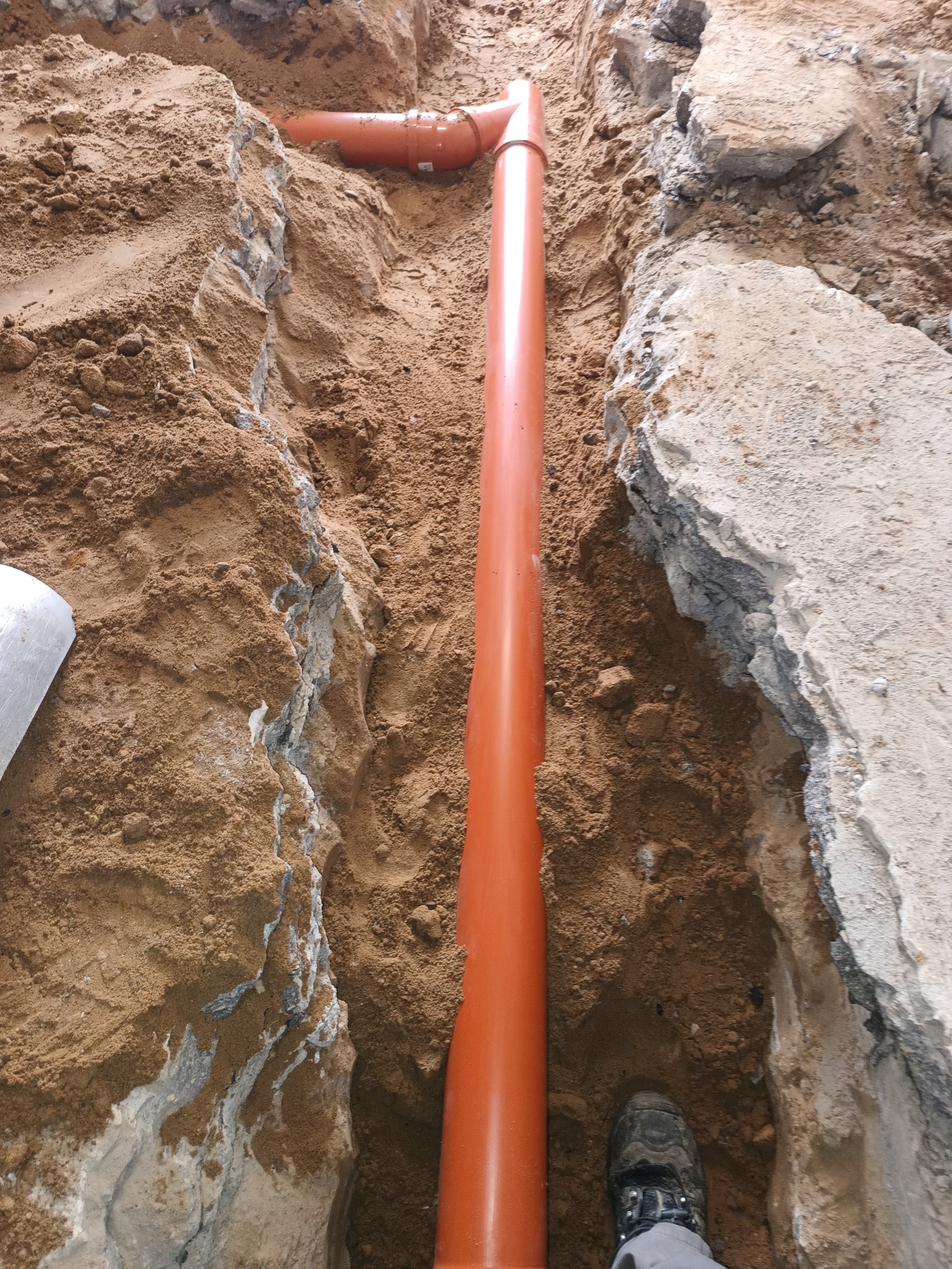 Pomarańczowa rura kanalizacyjna w wykopie ziemnym, widoczny fragment buta pracownika. Przyłącze wod-kan w trakcie realizacji, ziemia piaszczysta, fragmenty gruzu.