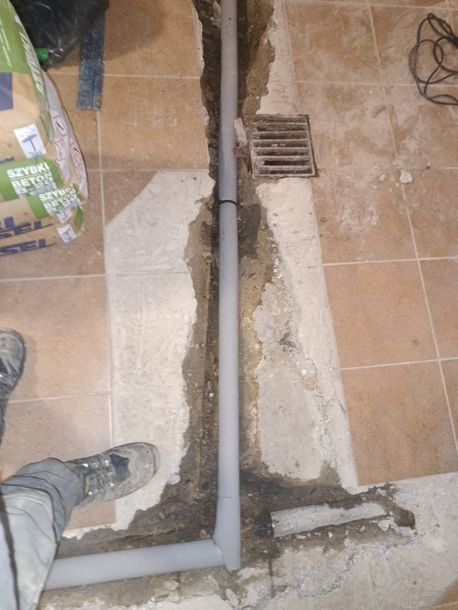 Szara rura kanalizacyjna w wykopanym bruzdzie w podłodze z płytek, widoczna krata ściekowa, worek z betonem i but. Etap prac podczas instalacji.