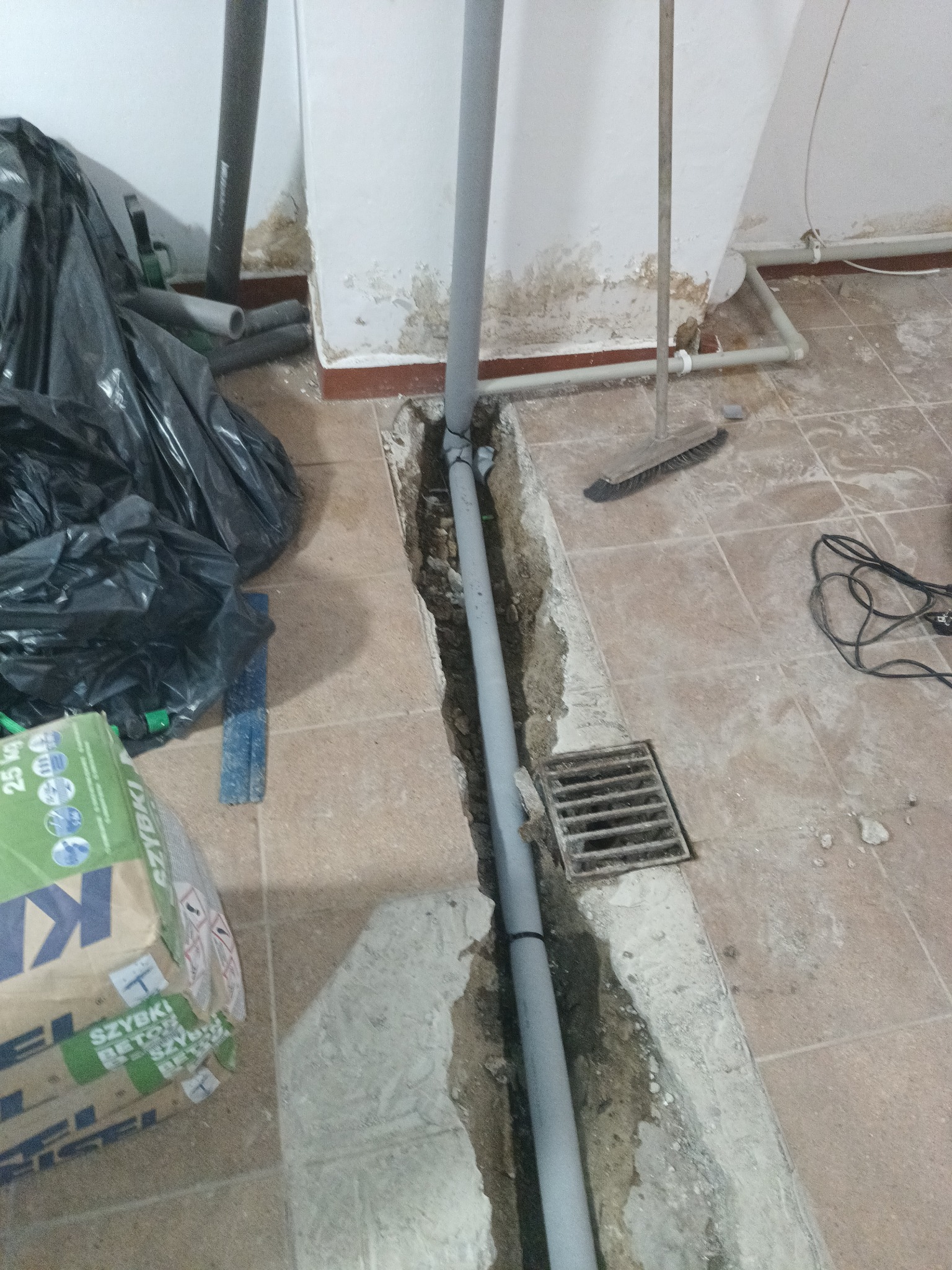 Szara rura kanalizacyjna w wykopie w podłodze z terakoty, obok kratka ściekowa, worek na śmieci, paczka betonu. Prace hydrauliczne w łazience.