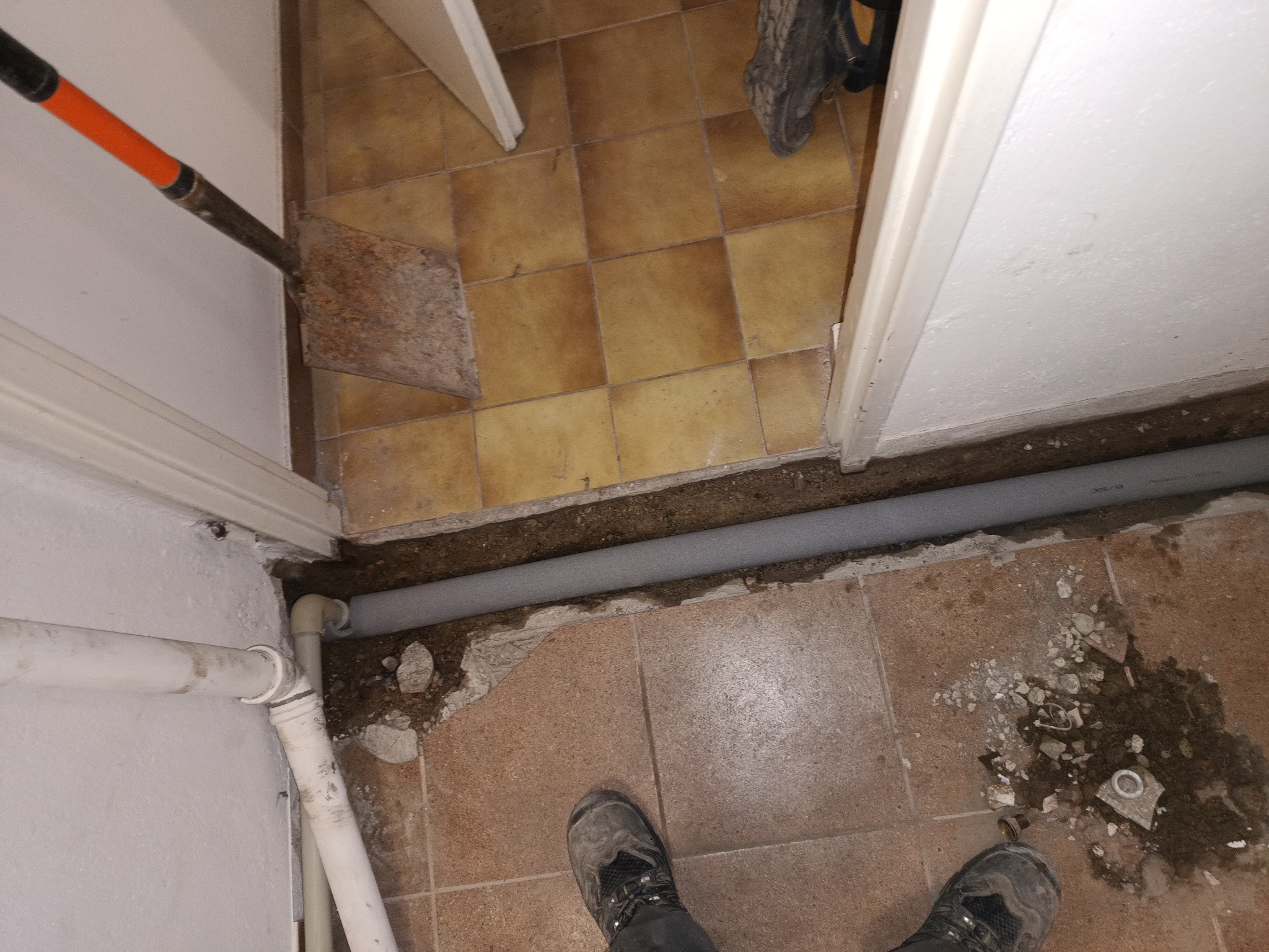 Szara rura kanalizacyjna w wykopanym rowku w podłodze z płytek, widoczne narzędzie i fragment buta. Prace hydrauliczne w trakcie remontu łazienki.