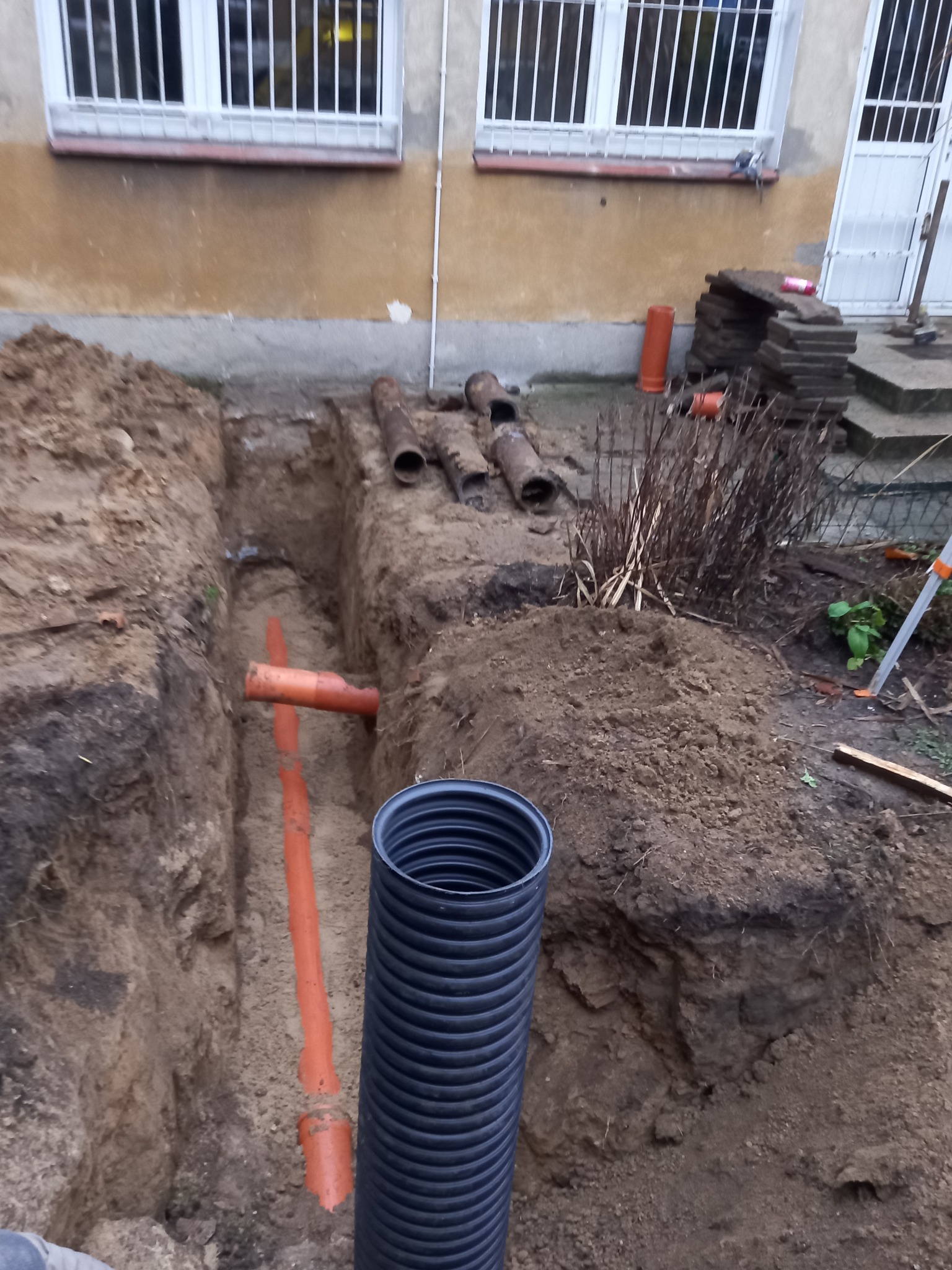 Wykop z pomarańczowymi rurami kanalizacyjnymi i czarną rurą karbowaną. Rury leżące na ziemi w tle. Prace ziemne przy budynku z oknami.