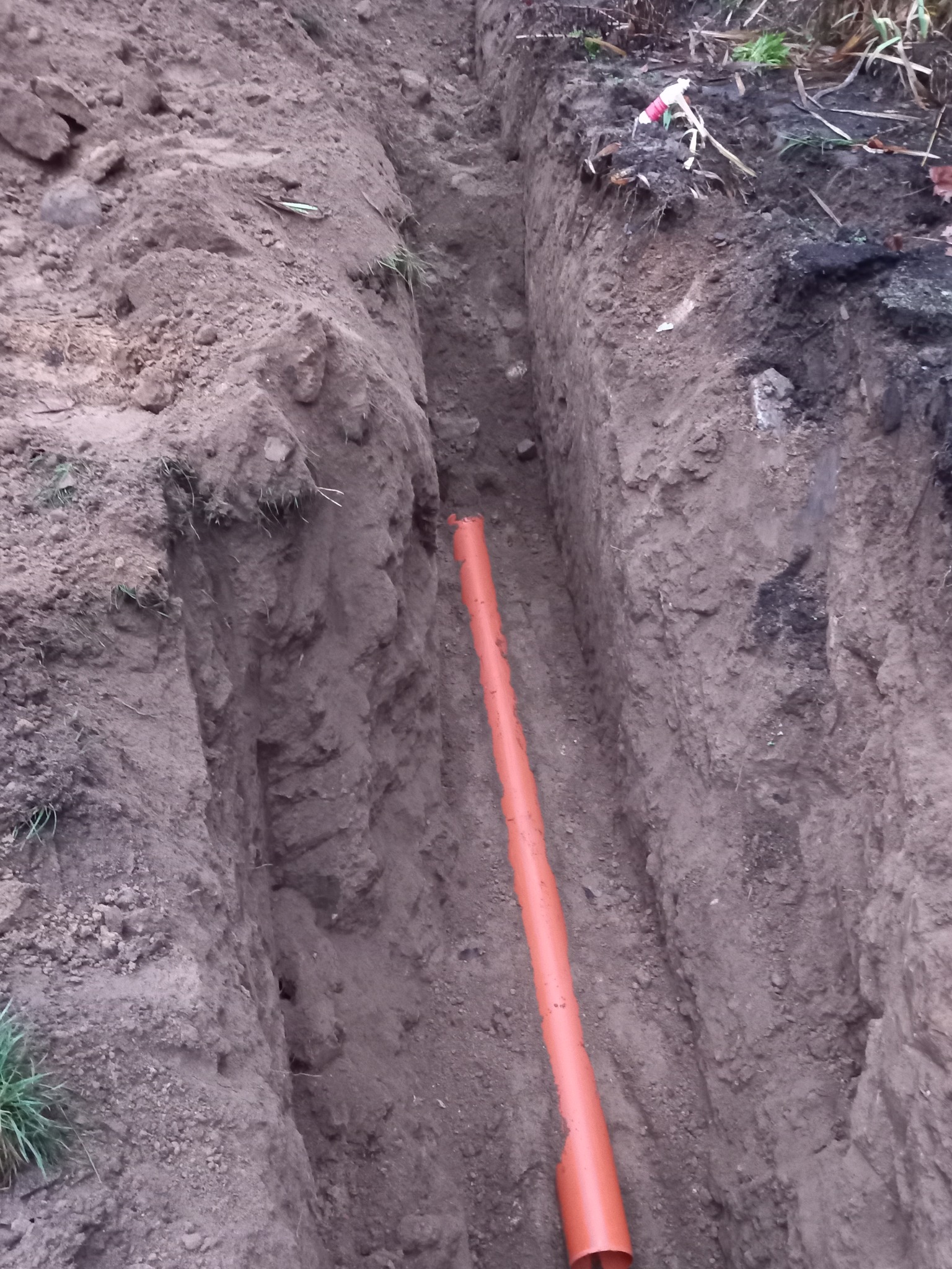 Pomarańczowa rura w wykopie ziemnym. Widok z góry na instalację kanalizacyjną w trakcie prac ziemnych, z widoczną rurą w wąskim wykopie.