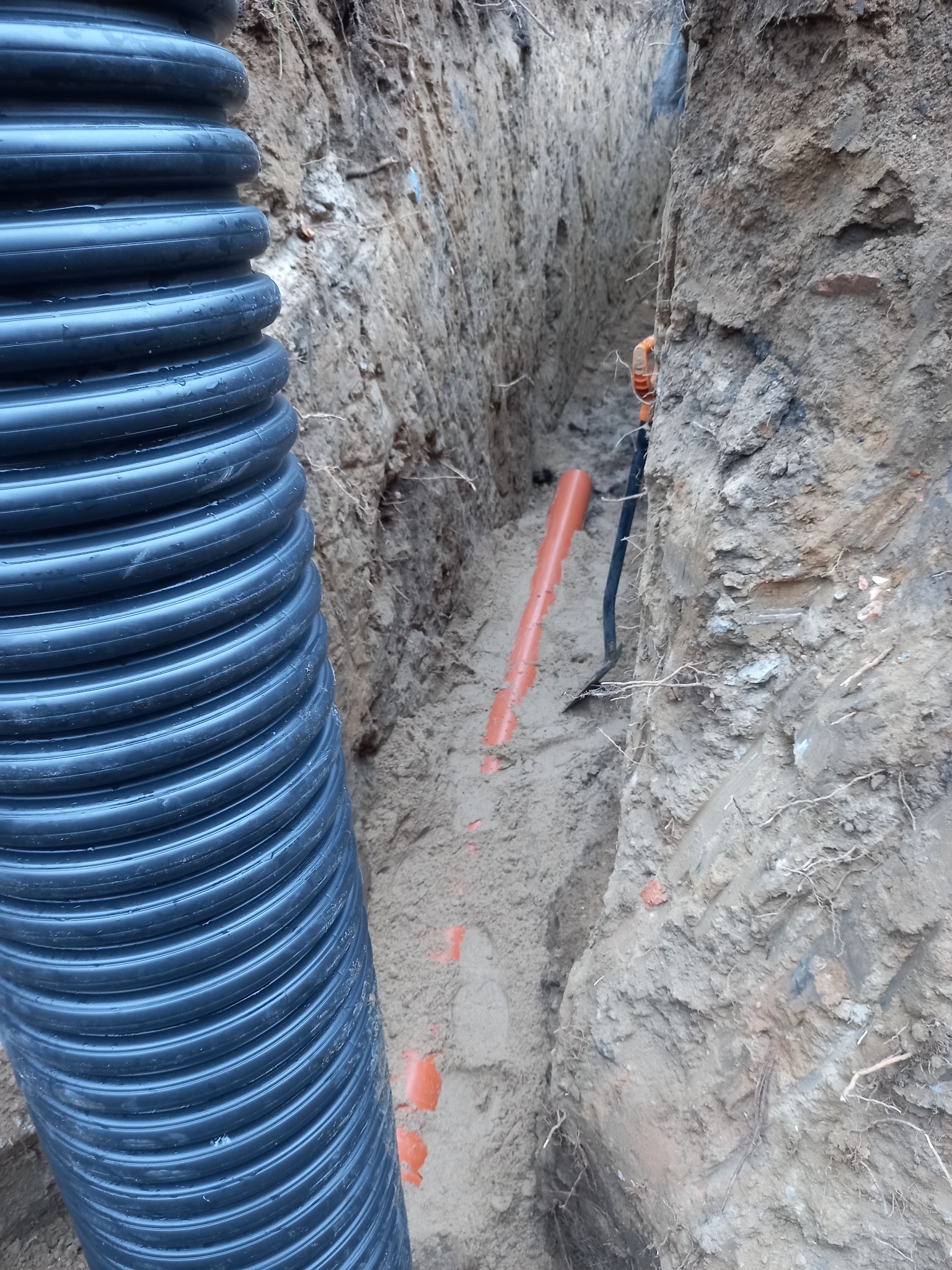 Wykop z ułożonymi rurami drenażowymi i pomarańczową rurą kanalizacyjną. Widoczna łopata w wykopie.