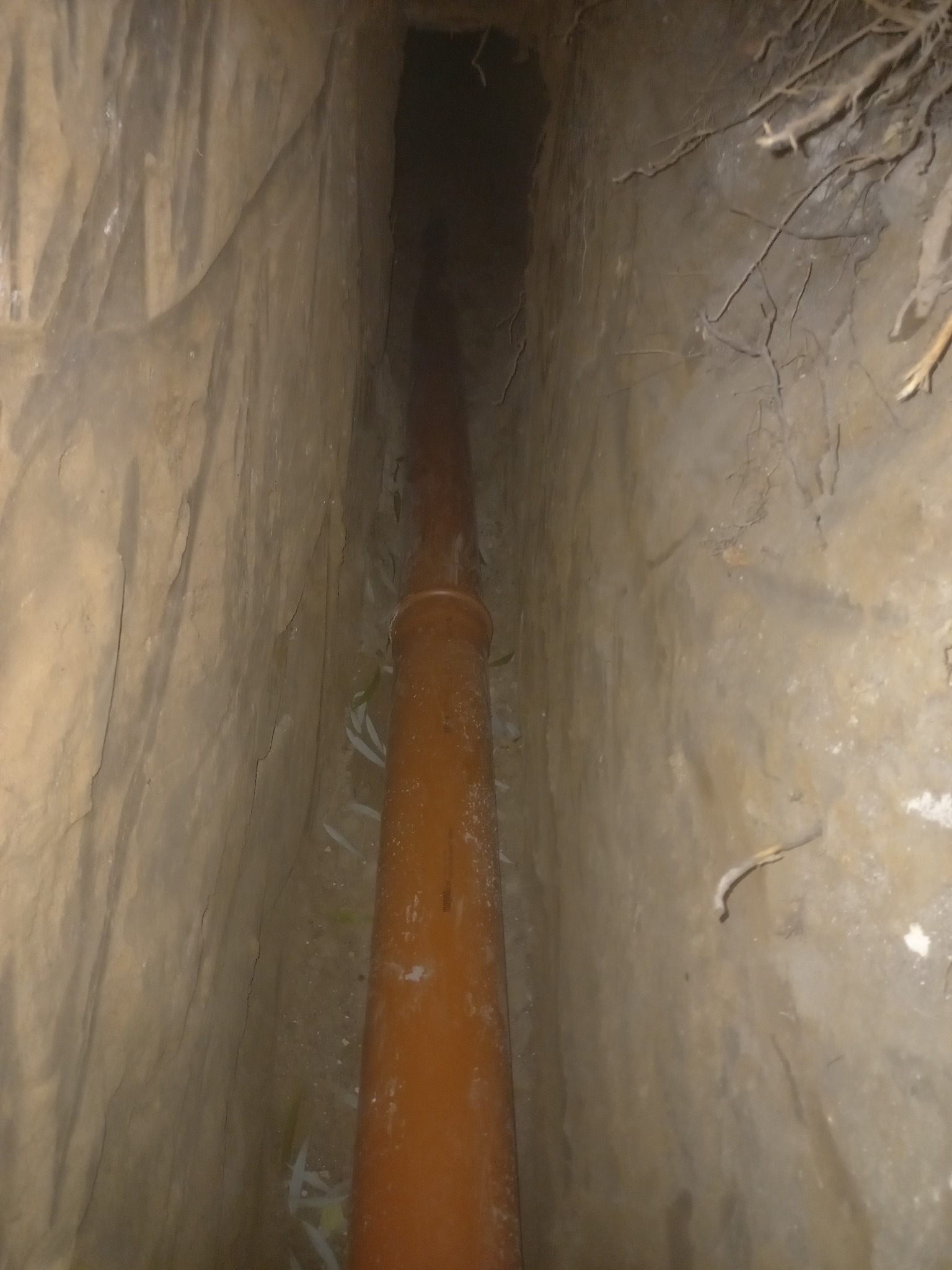 Brązowa rura kanalizacyjna ułożona w wykopie ziemnym, widok z góry. Ściany wykopu nierówne, z widocznymi śladami dłuta i korzeniami.