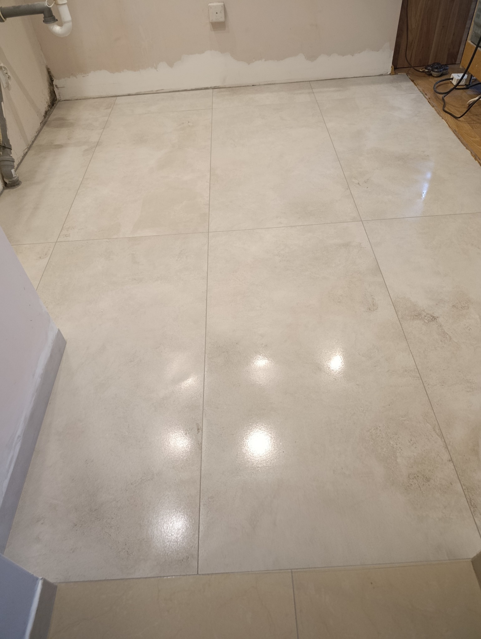 Ułożona, jasna podłoga z płytek w trakcie remontu łazienki. Widoczne niedokończone ściany i rury. Duże, kwadratowe płytki z delikatnym wzorem.