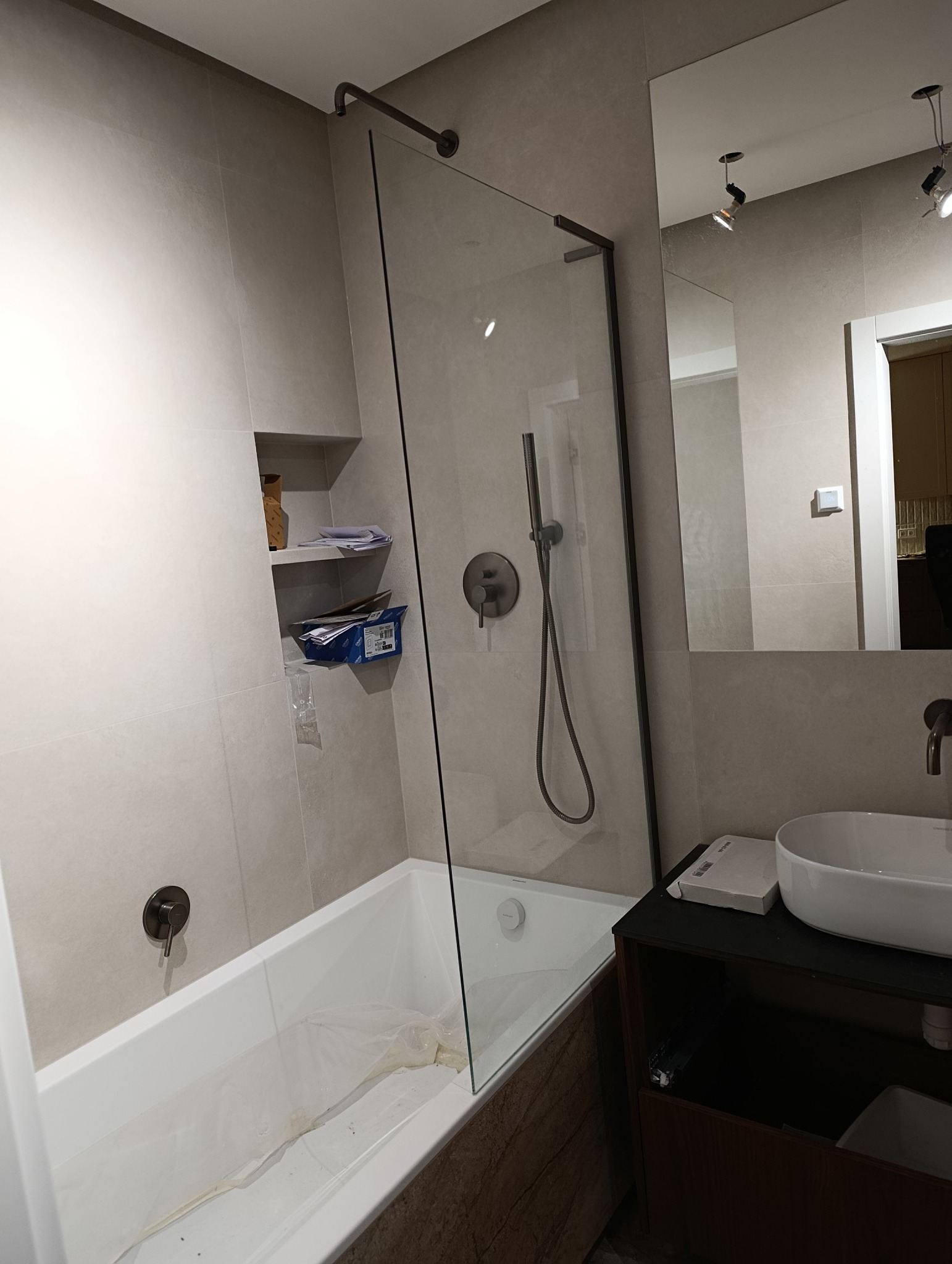 Nowoczesna łazienka z wanną, szklanym parawanem i umywalką nablatową. Beżowe płytki, minimalistyczna armatura w kolorze grafitu. Półka w ścianie z akcesoriami.