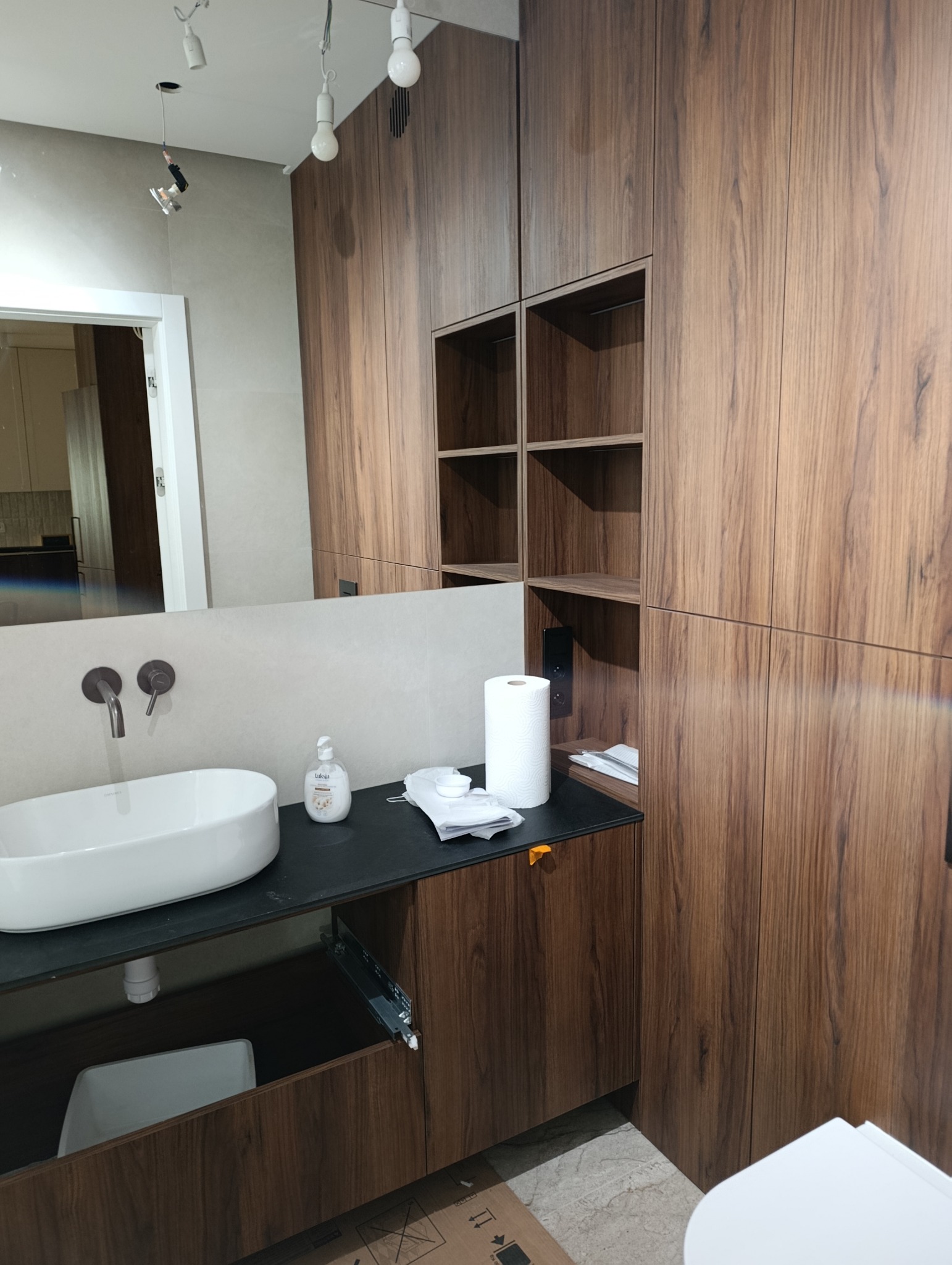 Nowoczesna łazienka z umywalką nablatową, szafką z drewnopodobnym fornirem i otwartymi półkami. Widoczne detale wykończenia i oświetlenie.