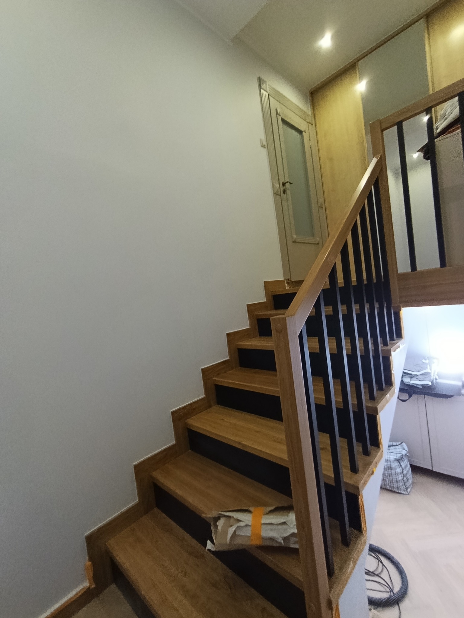 Drewniane schody z czarną balustradą w jasnym wnętrzu. Widoczna ściana, drzwi i fragment szafy. Ujęcie z dołu, podkreślające detale wykończenia.