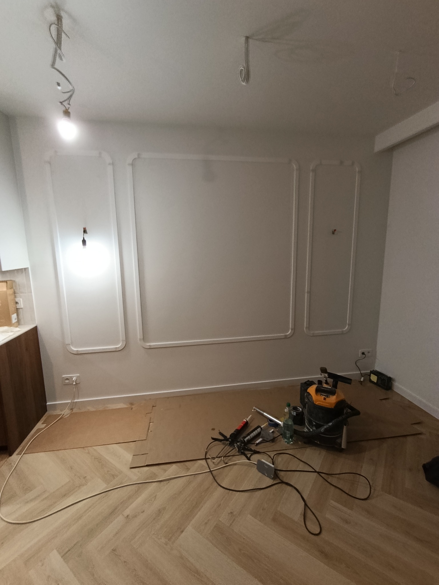 Białe listwy dekoracyjne na ścianie w trakcie montażu. Na podłodze parkiet w jodełkę, narzędzia i odkurzacz. Widoczne niedokończone oświetlenie sufitowe.