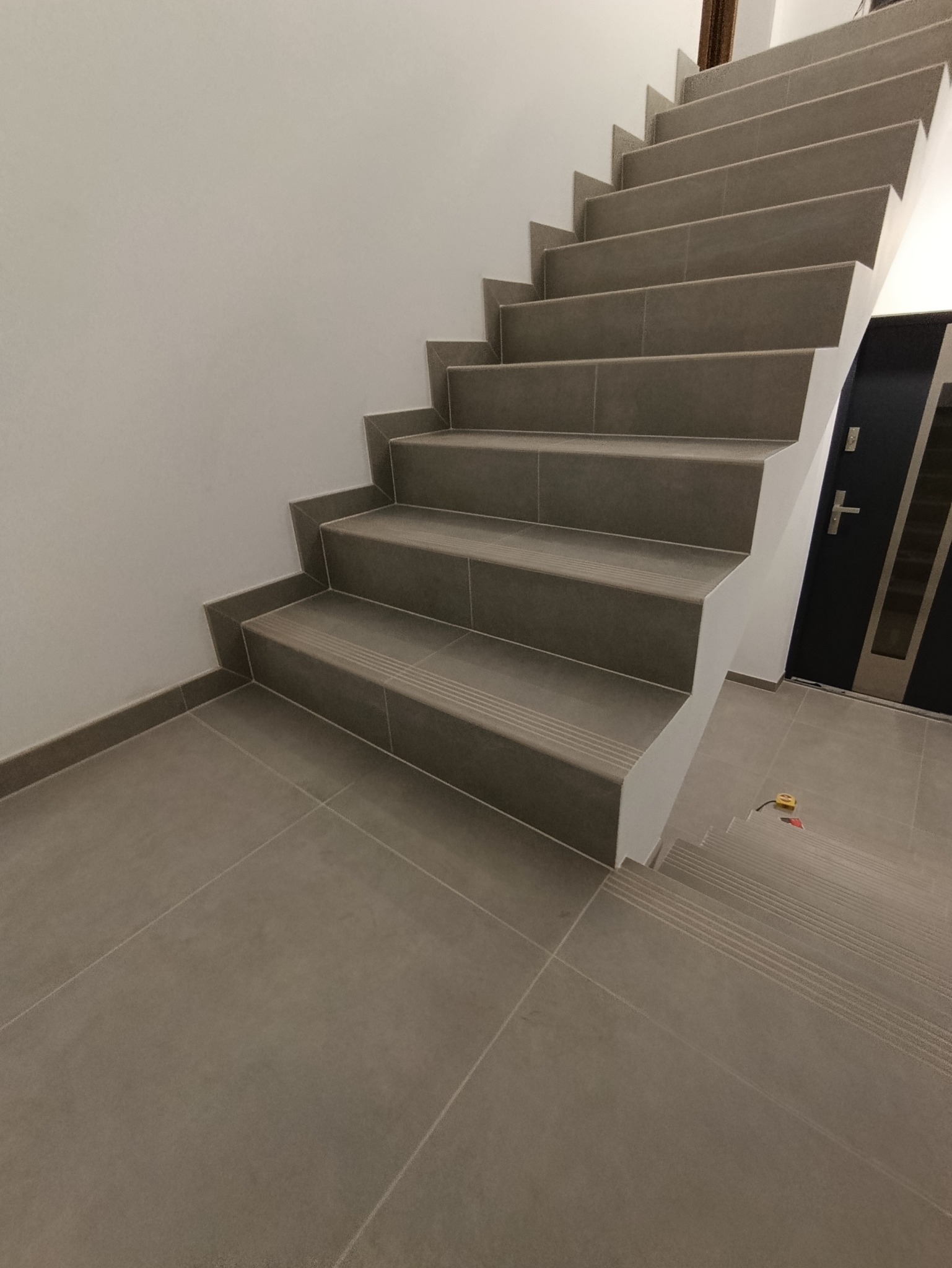 Wykończone schody z szarych płytek z antypoślizgowymi paskami, prowadzące w górę. Widoczna podłoga z płytek i fragment drzwi wejściowych. Minimalistyczny design.