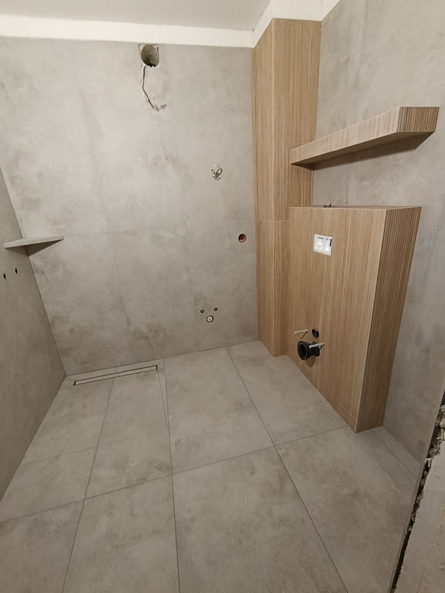 Wnętrze łazienki w trakcie wykończenia: szare płytki podłogowe i ścienne, zabudowa WC z drewnopodobną okładziną, widoczne instalacje. Minimalistyczny design.