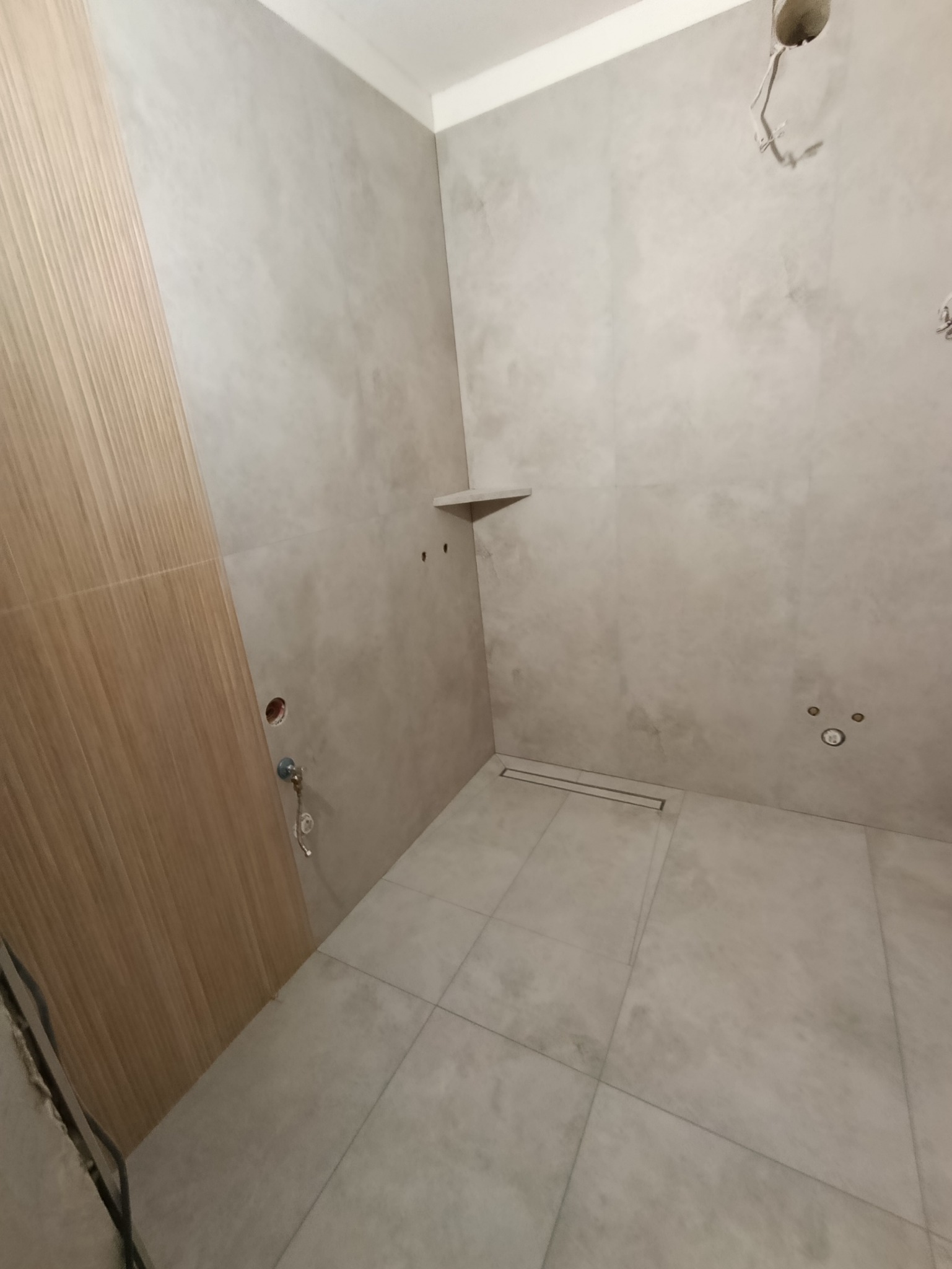 Wykończona łazienka z szarymi płytkami, drewnianą ścianą i odpływem liniowym. Widoczne przygotowane punkty instalacyjne i półka. Minimalistyczny design.