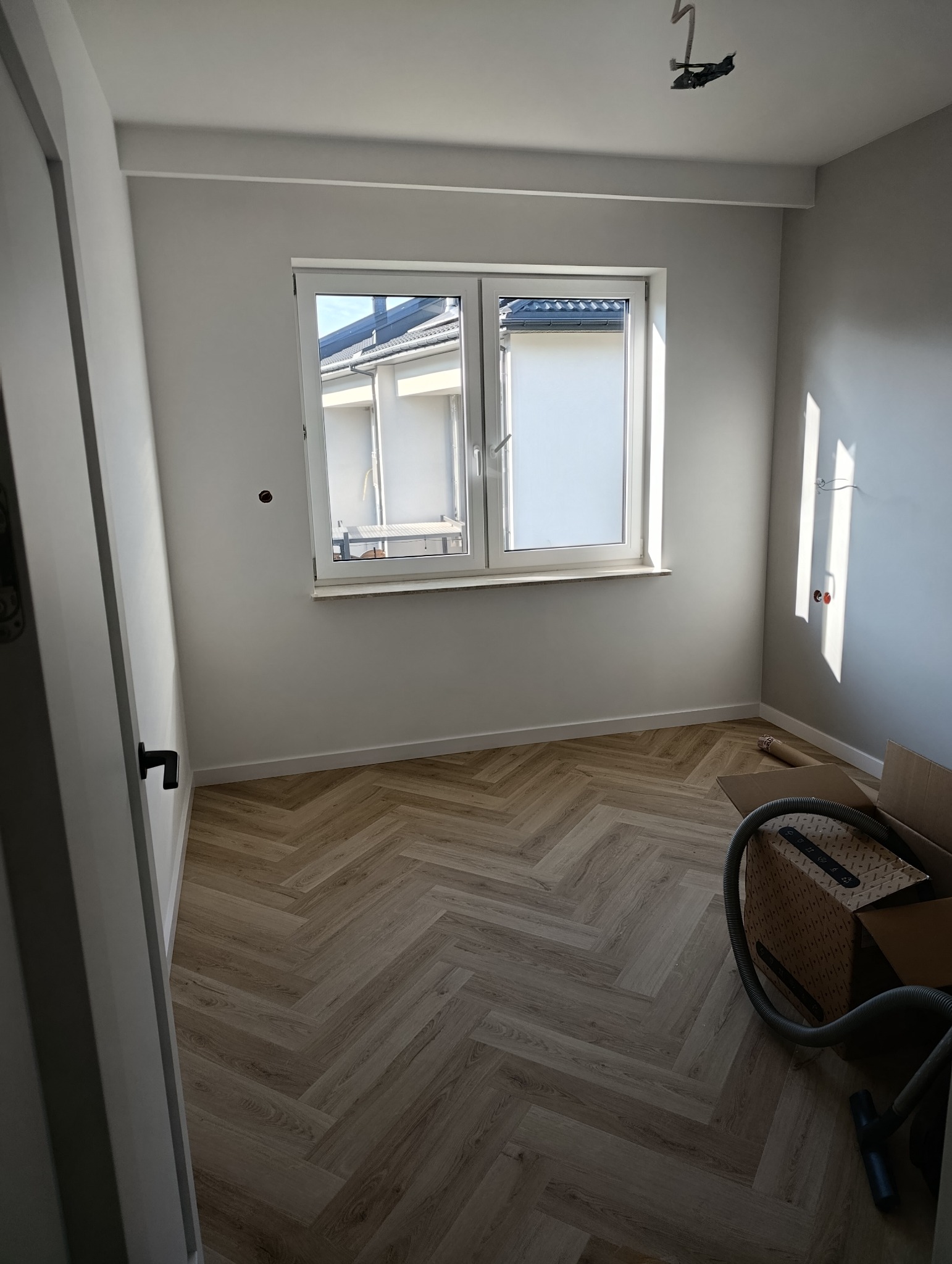 Jasny pokój po remoncie z parkietem ułożonym w jodełkę. Widoczne okno, białe ściany i fragment odkurzacza. Minimalistyczne wykończenie wnętrza.