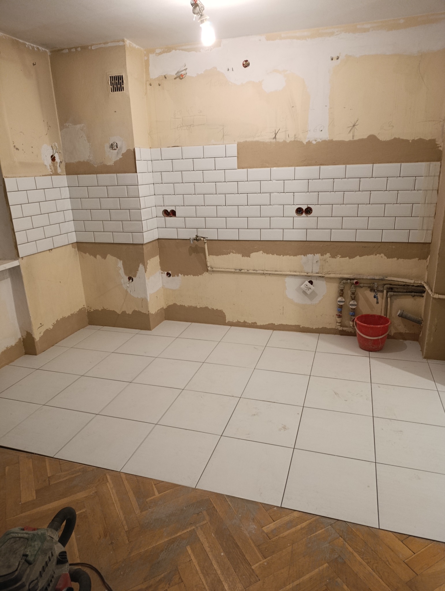 Remont kuchni w trakcie: białe płytki na ścianach i podłodze, widoczne instalacje, ślady po starych płytkach. Na podłodze parkiet i narzędzie.