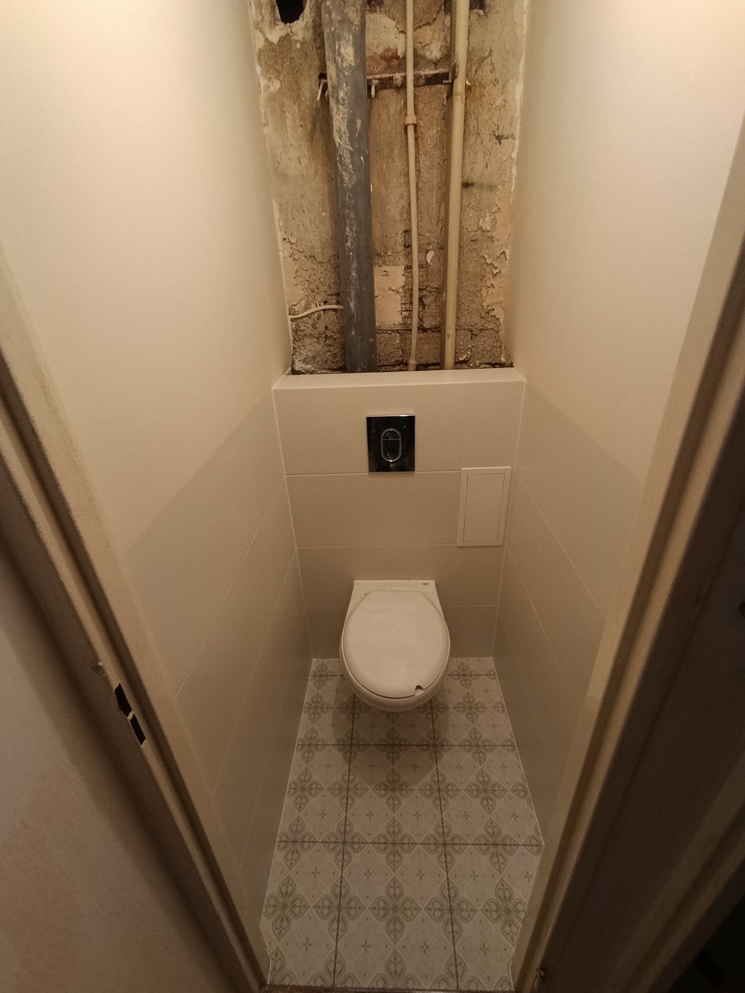 Mała toaleta po remoncie: białe płytki ścienne, wzorzysta podłoga, wisząca muszla. Widoczne rury na suficie przed zabudową. Ujęcie z perspektywy wejścia.