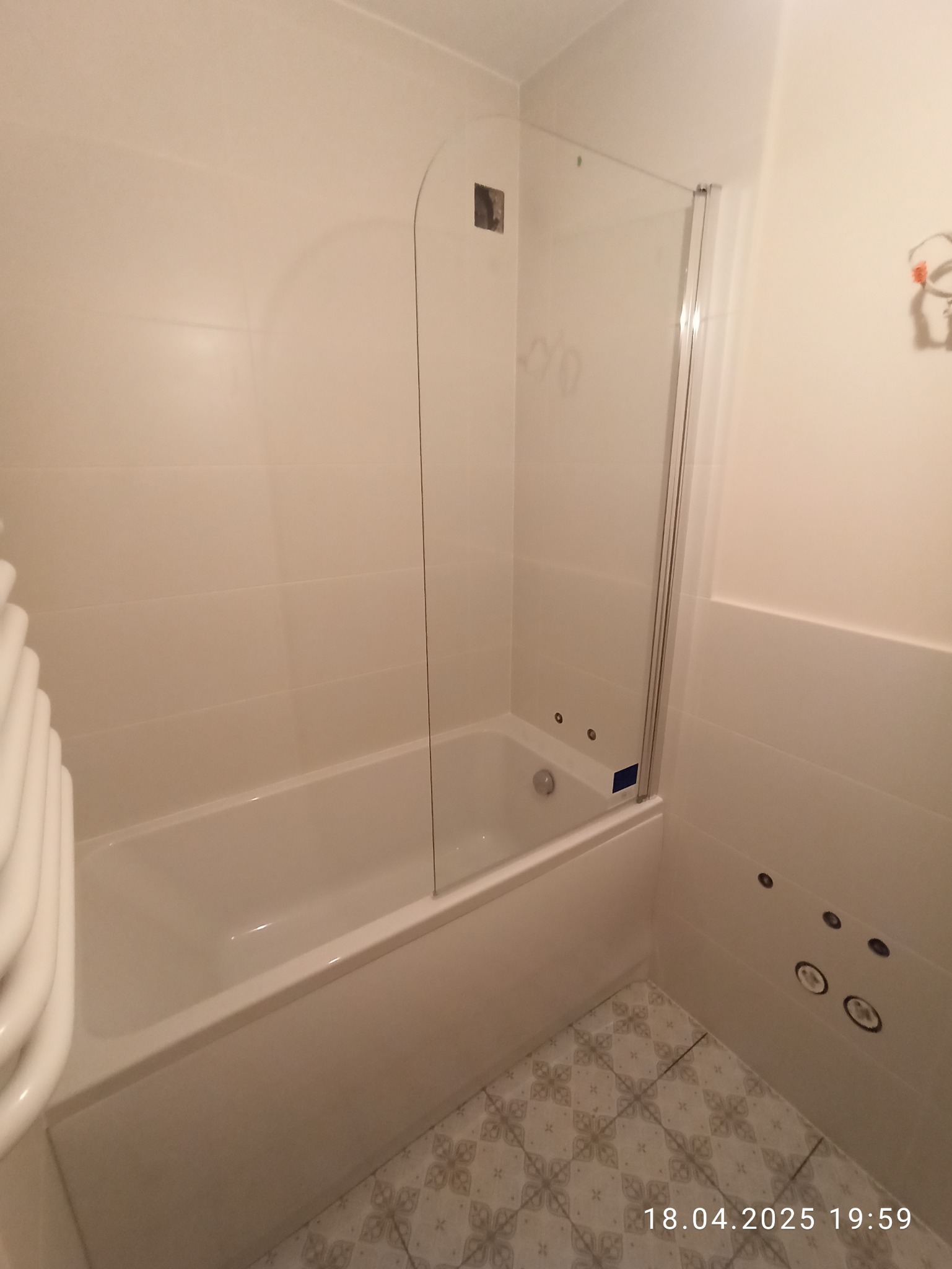 Biała wanna z hydromasażem i szklanym parawanem nawannowym w wykafelkowanej łazience. Widoczne dysze hydromasażu i wzorzysta podłoga.