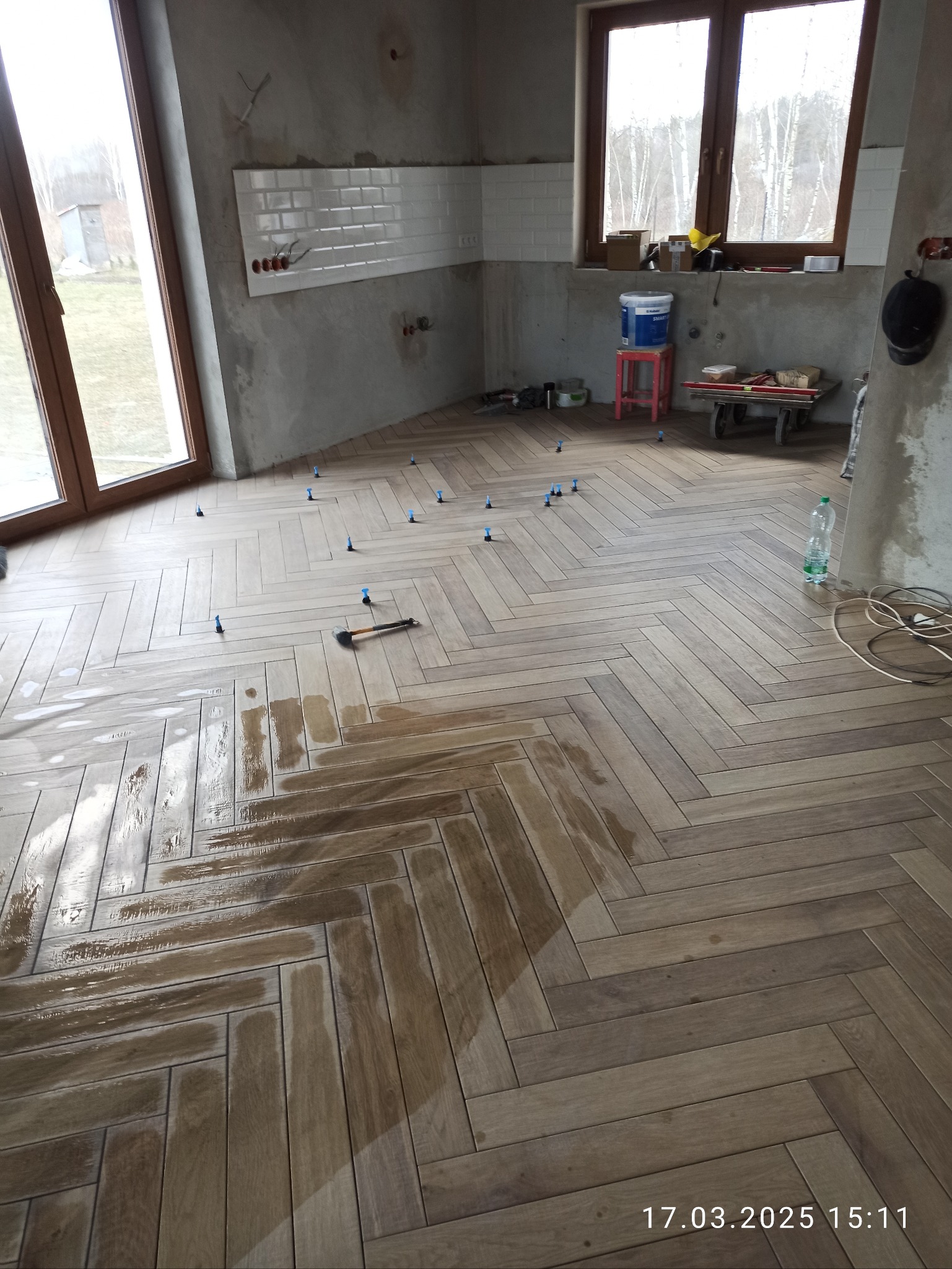 Układanie parkietu w jodełkę w trakcie prac. Widoczne płytki, poziomice, młotek i krzyżyki dystansowe. Częściowo położona glazura na ścianie. Surowy stan wykończenia.