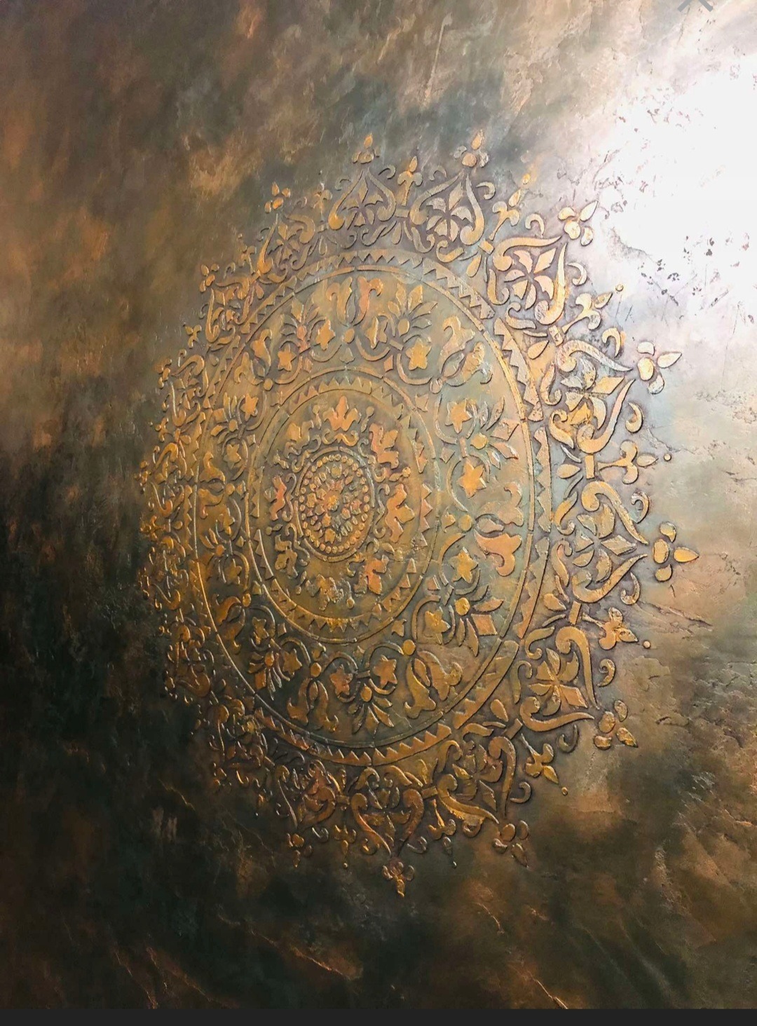 Dekoracyjna ściana z efektem metalicznego tynku strukturalnego i złocistym, wypukłym ornamentem w kształcie mandali, oświetlona z prawej strony.