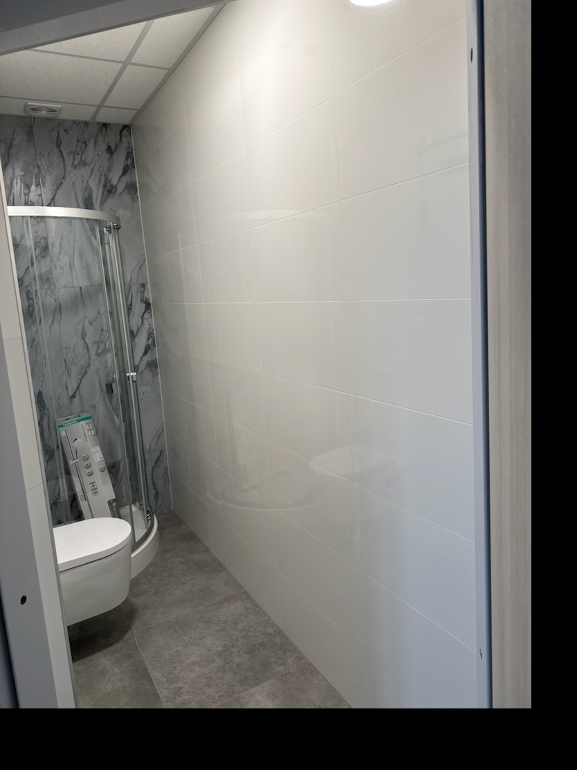 Wykończona łazienka z kabiną prysznicową i toaletą. Ściany wyłożone białymi, błyszczącymi płytkami. Na podłodze szare płytki. Widok zza framugi drzwi.