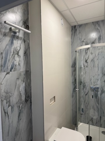 Wykończona łazienka z kabiną prysznicową i marmurowymi płytkami. Na ścianie designerska lampa nad toaletą. Widoczne białe płytki i sufit podwieszany.