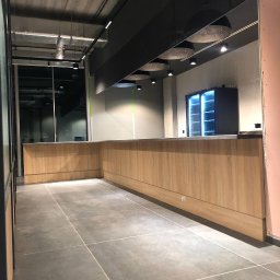 ARTmeble - Długa, drewniana lada w nowoczesnym wnętrzu komercyjnym z szarą podłogą i widoczną instalacją sufitową. W tle chłodziarka z oświetleniem.