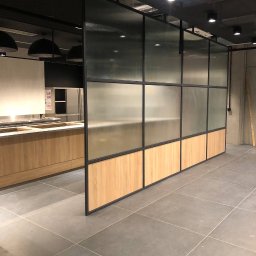 ARTmeble - Nowoczesna, szklana ścianka działowa z drewnianymi panelami w dolnej części, oddzielająca przestrzeń kuchenną. Widoczne oświetlenie sufitowe i fragment zabudowy kuchennej.