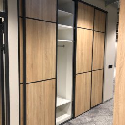 ARTmeble - Nowoczesna szafa przesuwna z drewnianymi frontami i czarnymi ramami, częściowo otwarta, ukazująca białe półki i drążek na ubrania. Widok z boku, szary dywan.