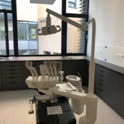 Szare szafki pod oknem w gabinecie dentystycznym, na pierwszym planie fotel dentystyczny z oprzyrządowaniem. Widok na budynek przez otwarte okno.