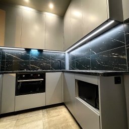 ARTmeble - Szara kuchnia z czarnym blatem i marmurowymi płytkami. Oświetlenie LED podkreśla nowoczesny design. Widoczny piekarnik i mikrofalówka.