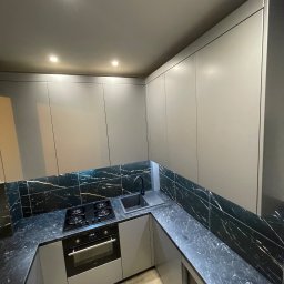 ARTmeble - Nowoczesna kuchnia z szarymi frontami i blatem z czarnego marmuru z białymi żyłami. Widoczna płyta grzewcza, piekarnik i zlew. Minimalistyczny design, oświetlenie punktowe.