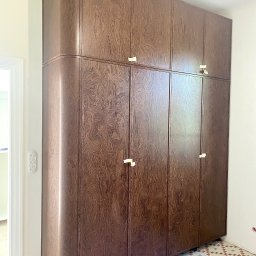 ARTmeble - Wysoka, brązowa szafa z zaokrąglonym bokiem i złotymi uchwytami, stojąca na kafelkach w stylu retro. Widoczny fragment ściany z włącznikami.