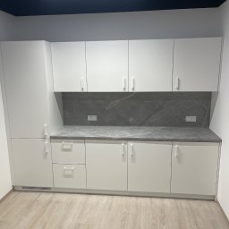 ARTmeble - Minimalistyczna, biała zabudowa kuchenna z szarym blatem i marmurowym panelem nad blatem. Widoczne gniazdka elektryczne. Jasna podłoga imitująca drewno.