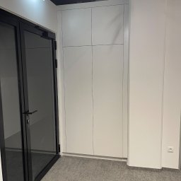 ARTmeble - Biała szafa wnękowa z czterema drzwiami, wkomponowana w ścianę, widoczna z perspektywy korytarza z szarym dywanem i czarnymi drzwiami.