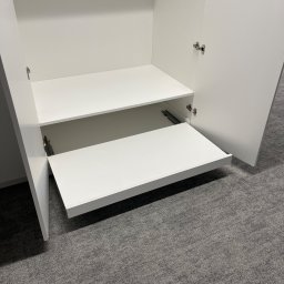 ARTmeble - Wnętrze białej szafy z wysuwaną półką na metalowych prowadnicach, widoczne zawiasy drzwi. Na podłodze szara wykładzina dywanowa.