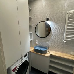 ARTmeble - Nowoczesna łazienka z jasną zabudową meblową, integrującą pralkę. Widoczna szafka z niebieską umywalką nablatową, okrągłe lustro, drewniany blat i faliste płytki ścienne.