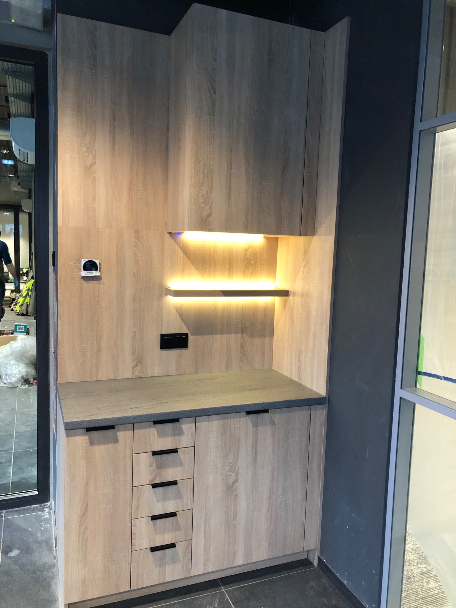 Jasna zabudowa meblowa z szufladami i podświetlanymi półkami, zamontowana w rogu pomieszczenia z dużym oknem. Widoczne detale wykończenia i oświetlenie led.