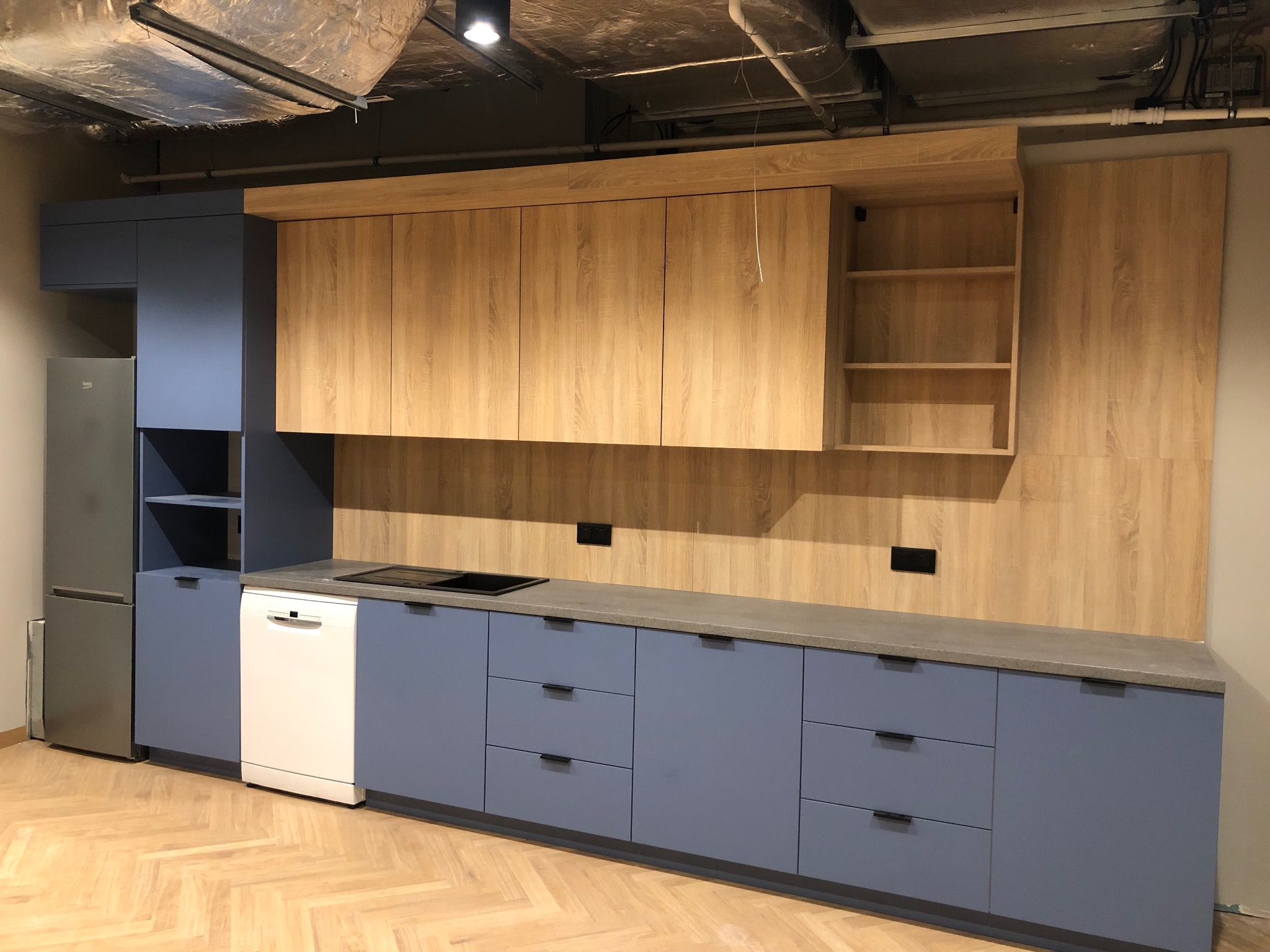 Nowoczesna kuchnia z drewnianymi szafkami górnymi i niebieskimi dolnymi, z blatem w kolorze betonu. Widoczna lodówka i płyta indukcyjna. Podłoga w jodełkę.