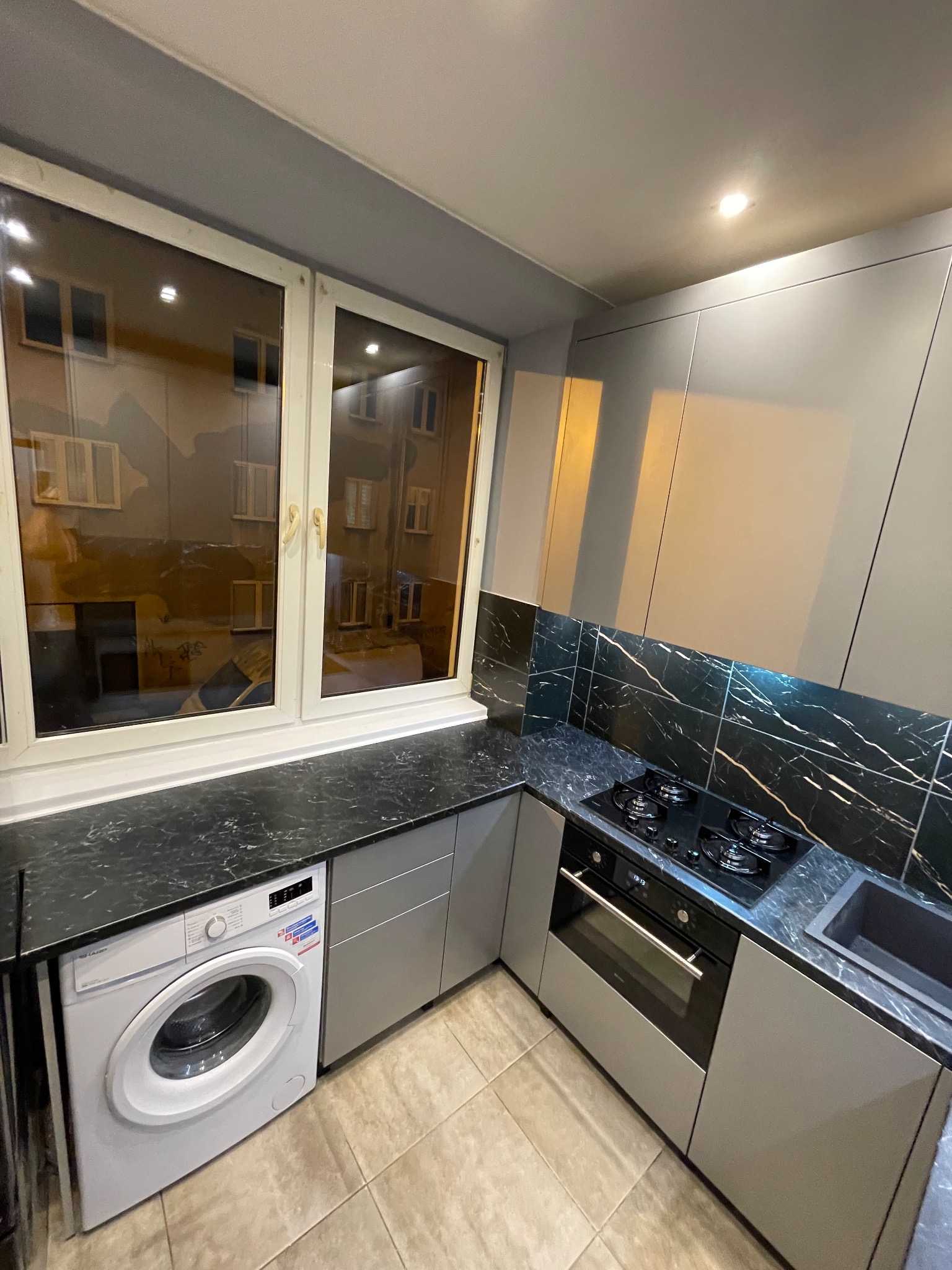 Szara kuchnia na wymiar z blatem imitującym marmur, zintegrowana z pralką pod oknem. Nowoczesne szafki bez uchwytów i płyta grzewcza z piekarnikiem.