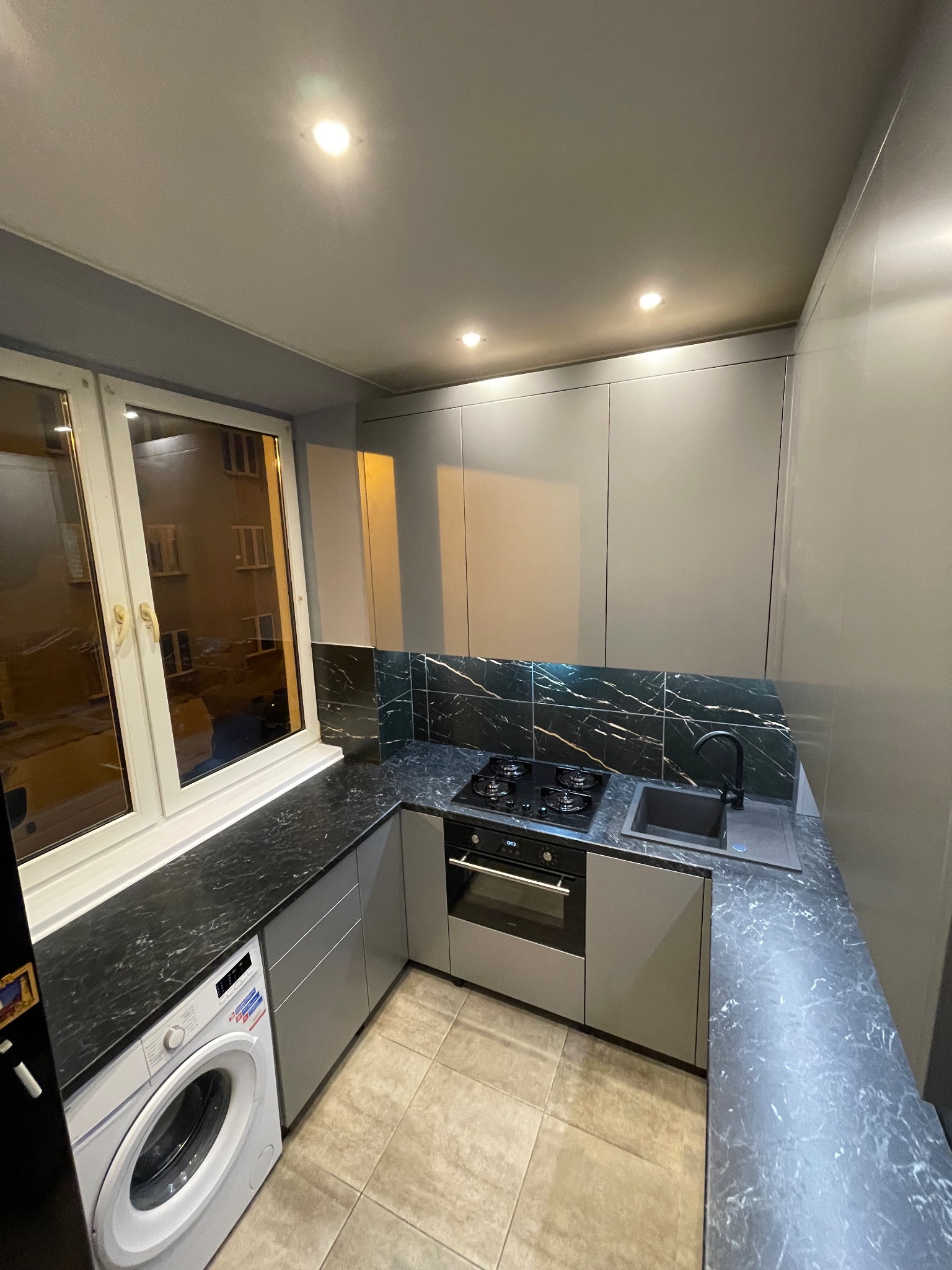 Szara, nowoczesna kuchnia z blatem imitującym marmur, zintegrowana z pralką pod oknem. Widok na płytę grzewczą i zlewozmywak. Oświetlenie punktowe.