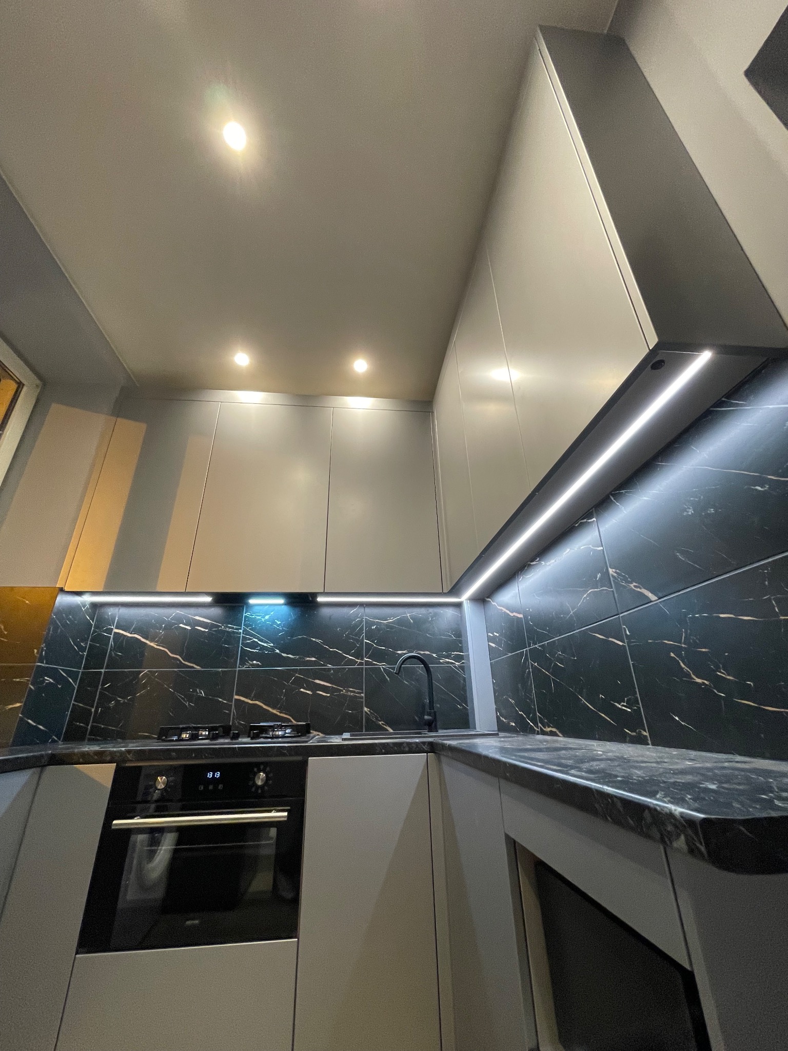 Nowoczesna kuchnia z szarymi frontami, czarnym blatem i marmurową ścianą z podświetleniem LED. Widok z dołu, podkreślający geometrię i oświetlenie.