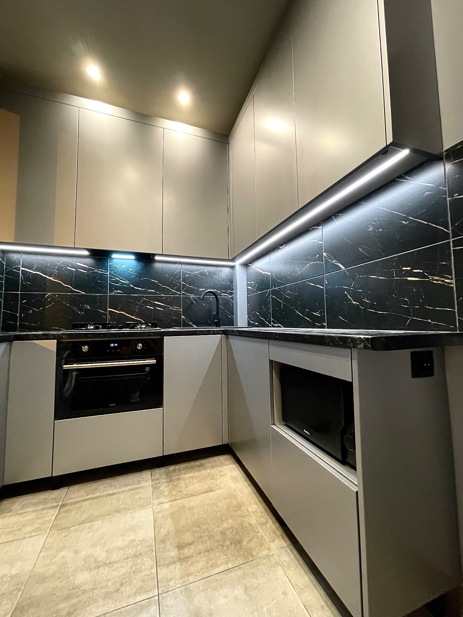 Szara kuchnia z czarnym blatem i marmurowymi płytkami. Oświetlenie LED podkreśla nowoczesny design. Widoczny piekarnik i mikrofalówka.