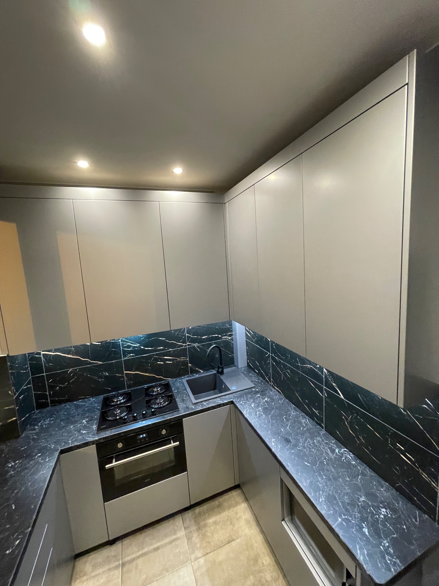 Nowoczesna kuchnia z szarymi frontami i blatem z czarnego marmuru z białymi żyłami. Widoczna płyta grzewcza, piekarnik i zlew. Minimalistyczny design, oświetlenie punktowe.