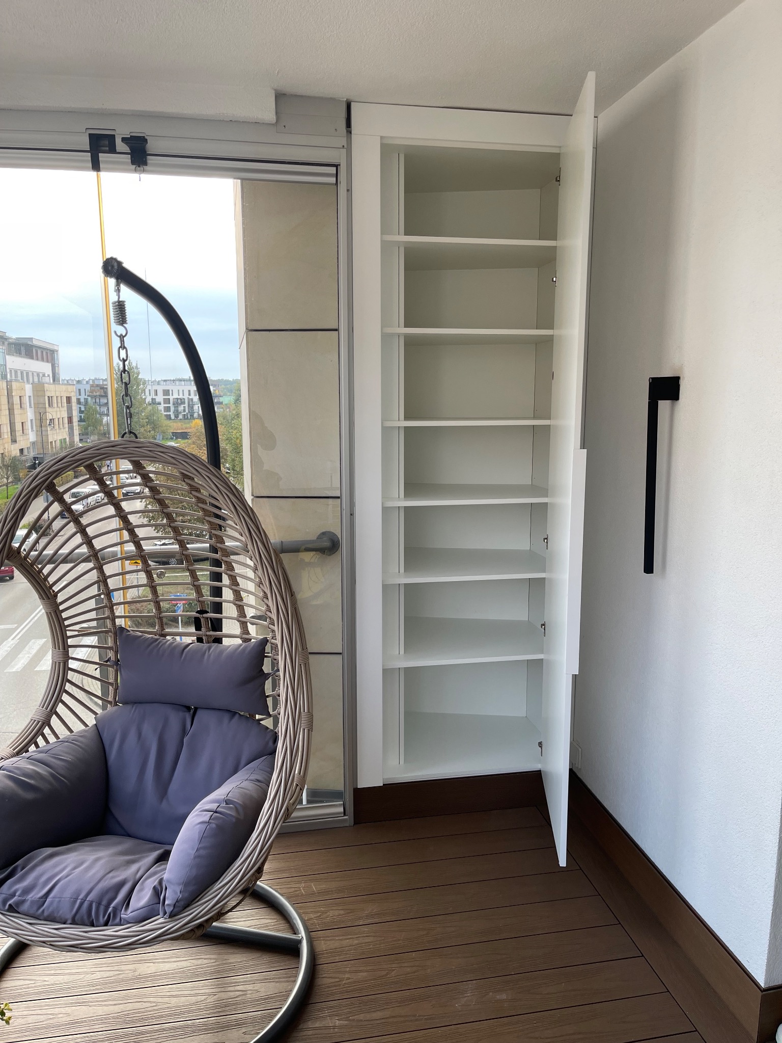 Biała szafa wnękowa z otwartymi drzwiami na balkonie z drewnianą podłogą i wiszącym fotelem. Widok na miasto w tle.
