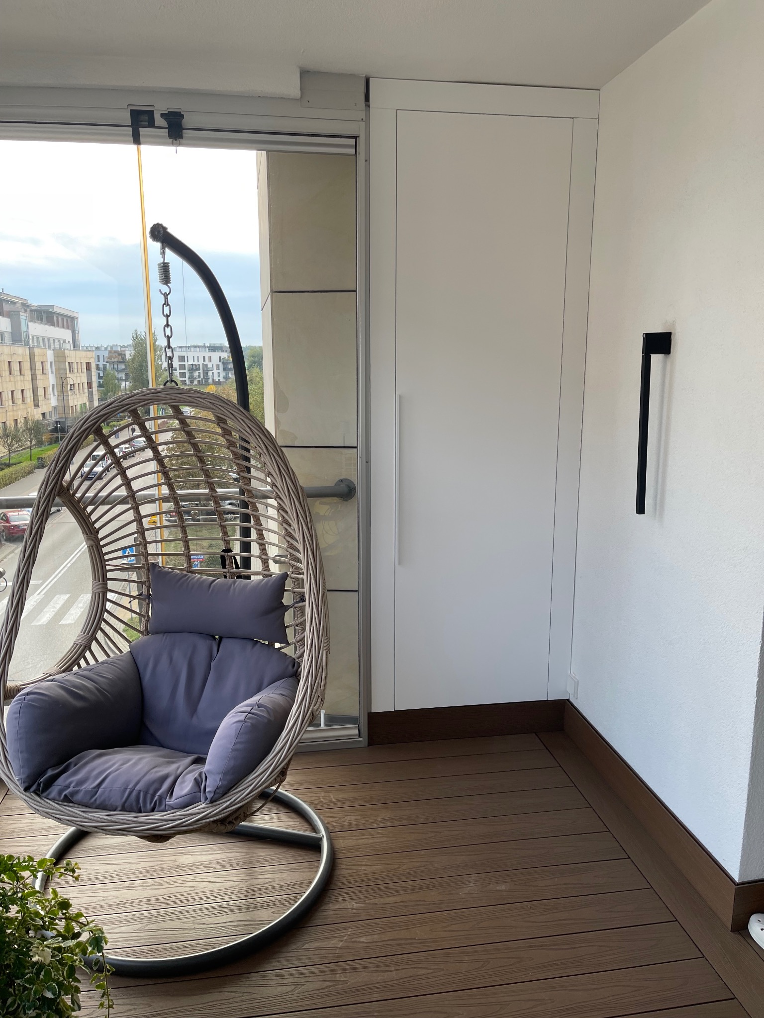 Balkon z zabudową szafy wnękowej i wiszącym fotelem. Drewniana podłoga, minimalistyczna lampa ścienna. Widok na miasto zza fotela.